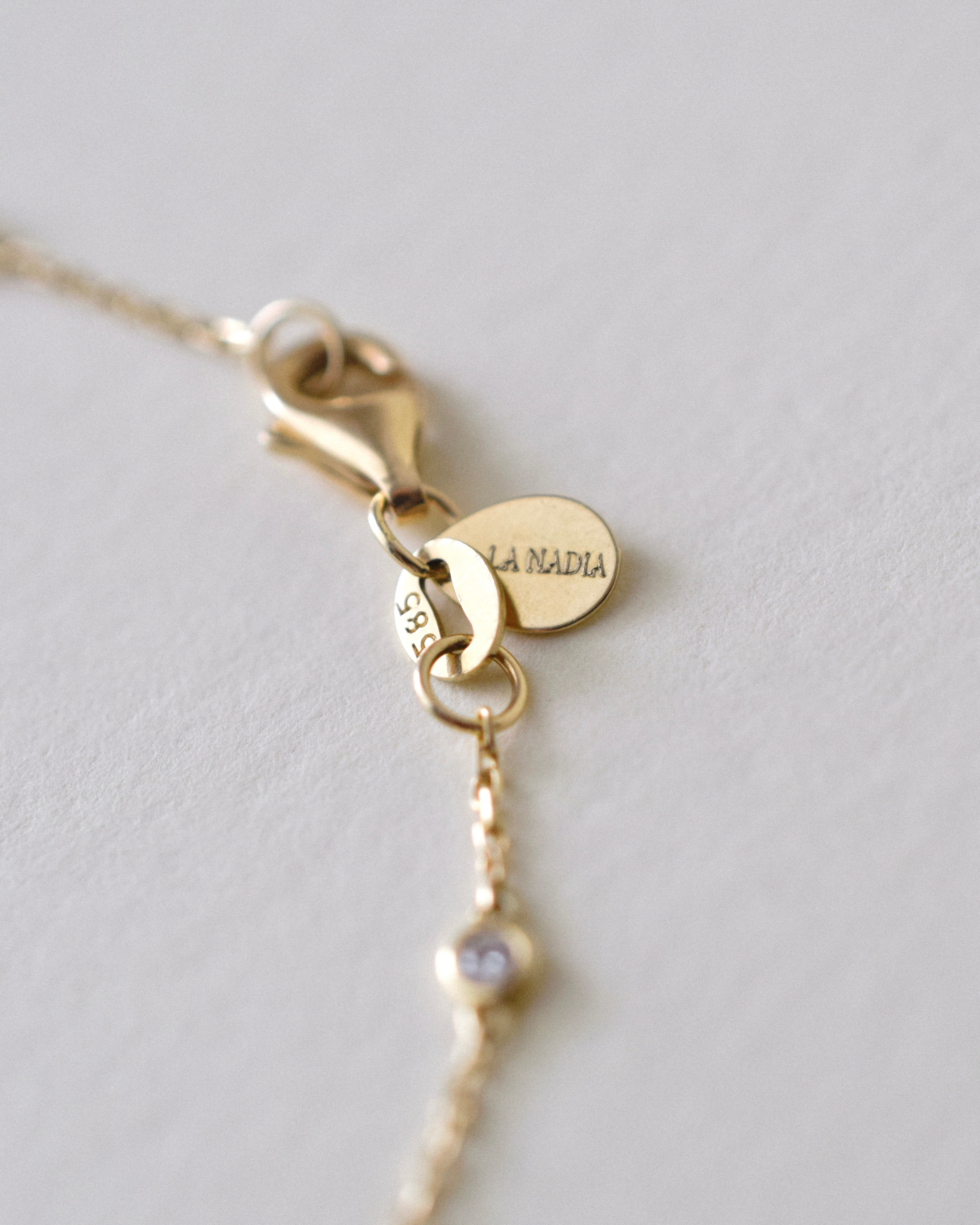 Gold necklace clasp with an oval-shaped jewelry tag with 'LA NADIA' engraving on a light gray background