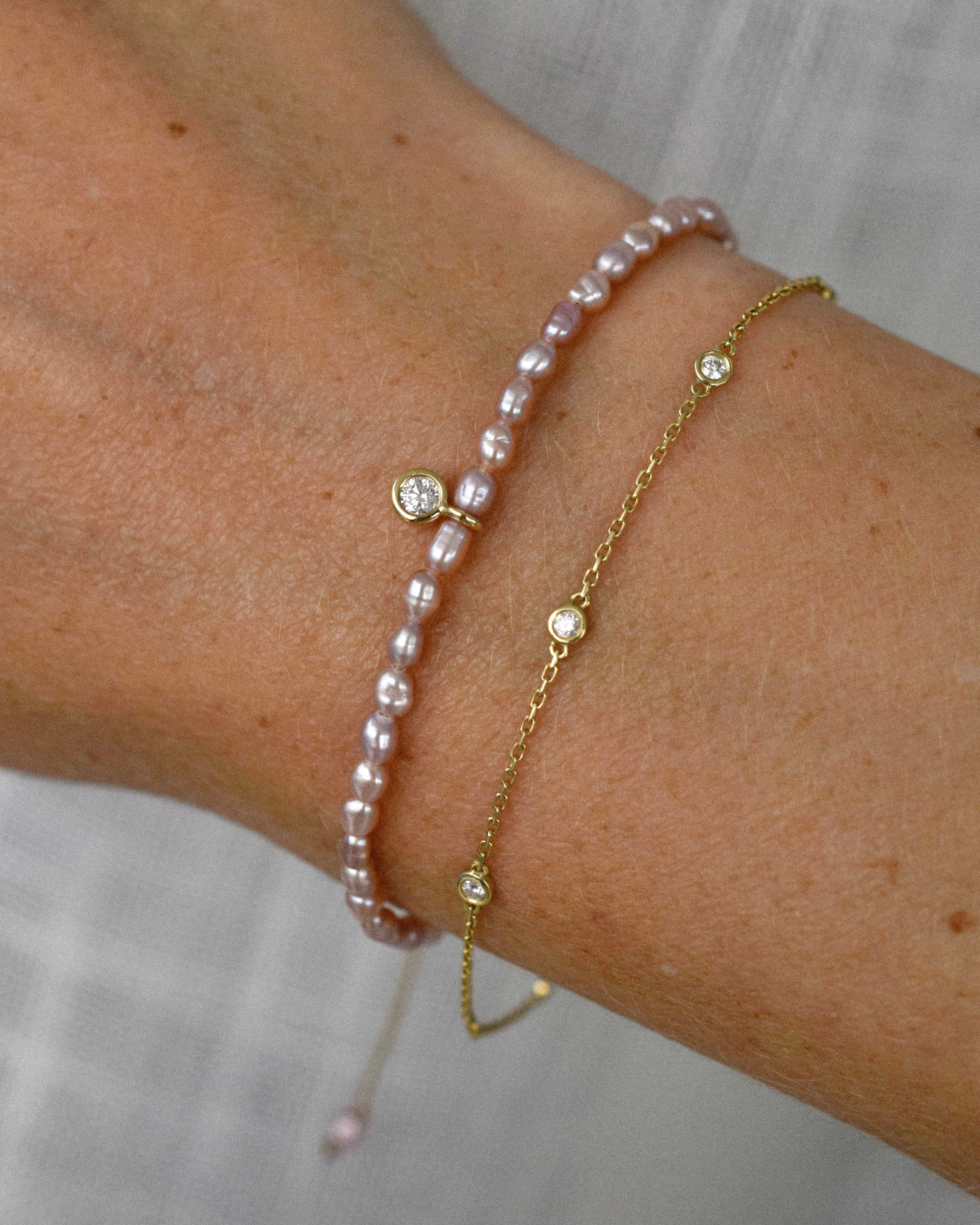 Close-up of a wrist wearing a purple pearl bracelet and a golden chain charm bracelet on a neutral background