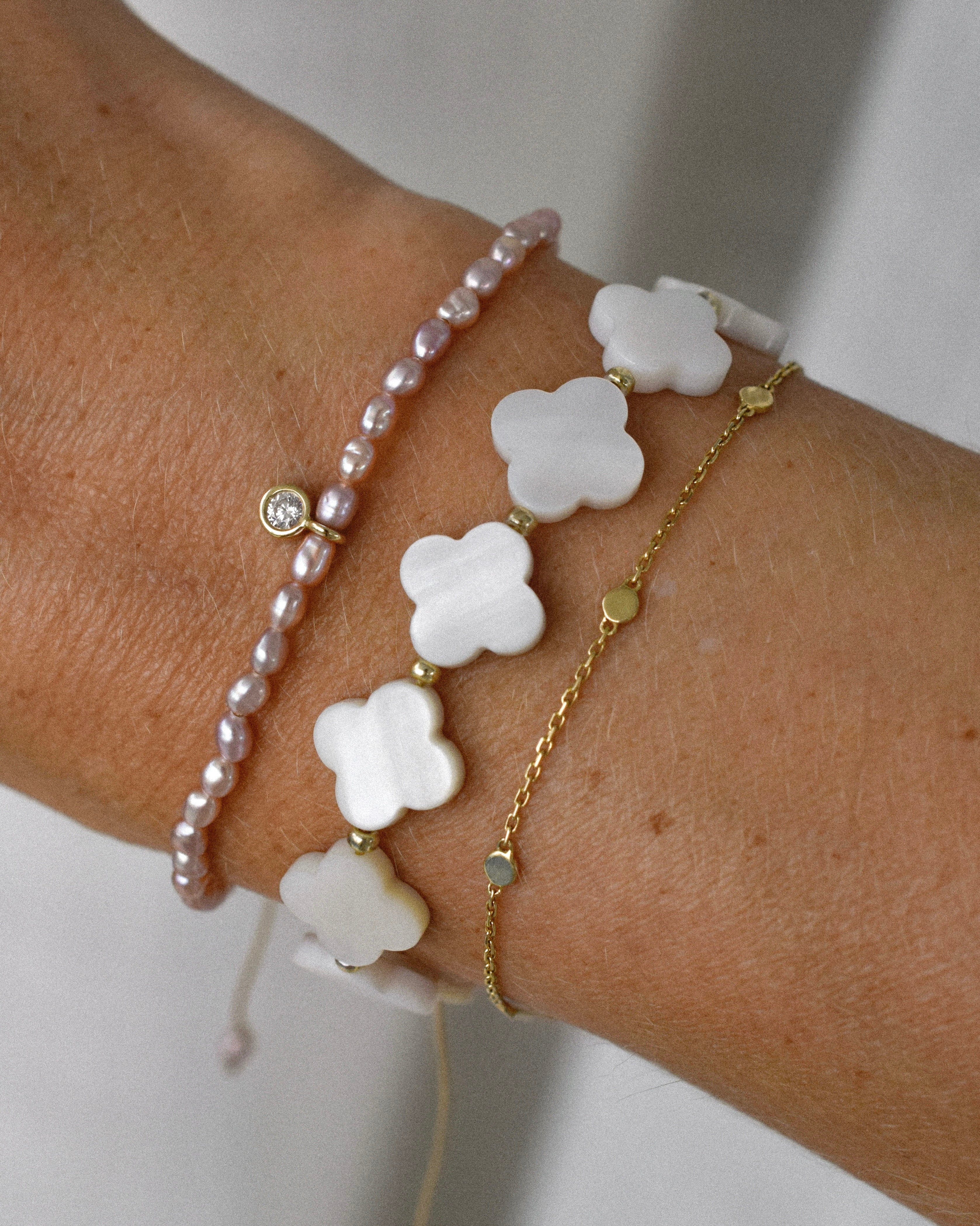 Close-up of a wrist wearing multiple bracelets stacked with clover and pearl designs on a neutral background