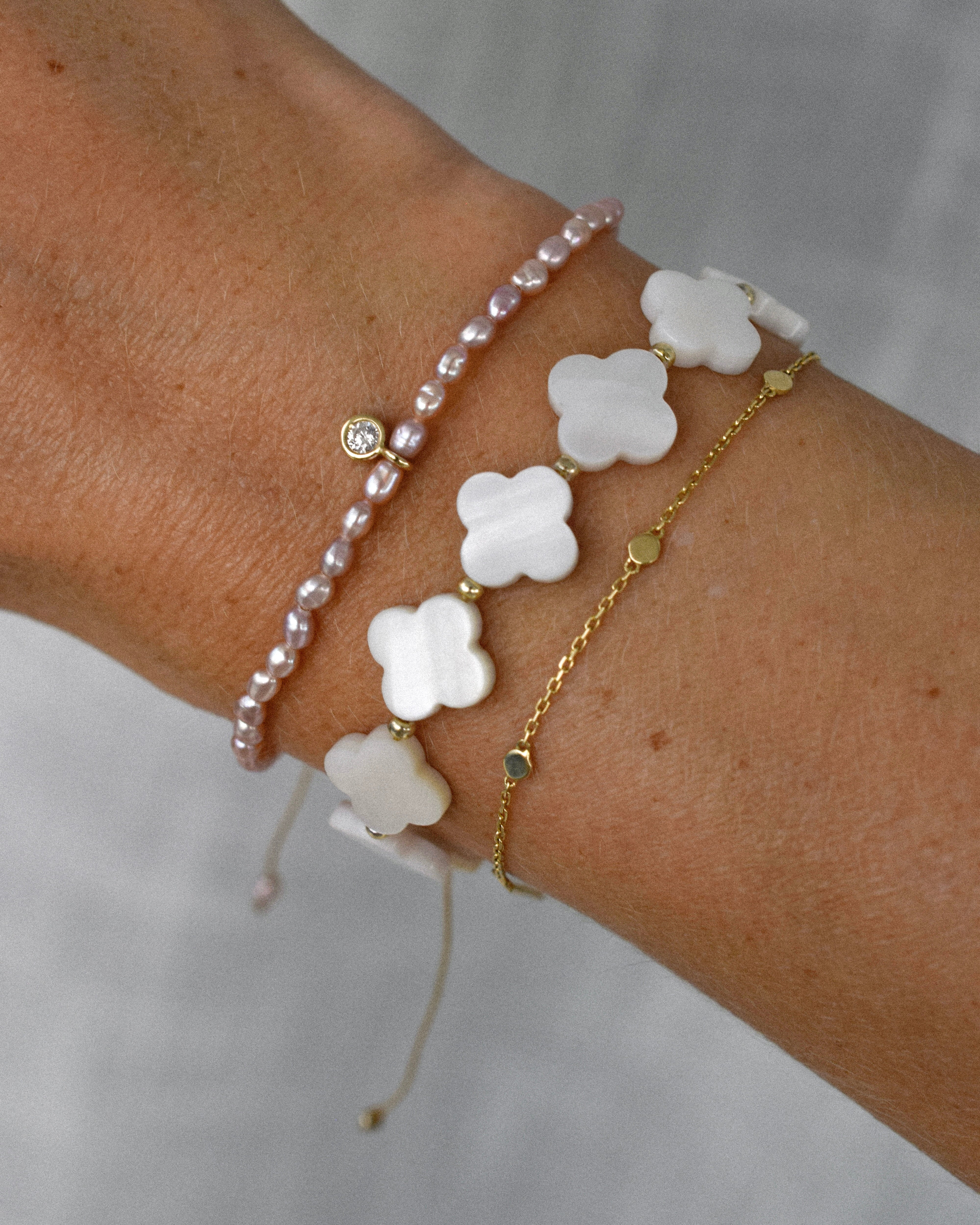 Pracelet stack with clover-shaped mother of pearl beads, purple freshwater pearls and a diamond solitaire charm on a wrist against a neutral background