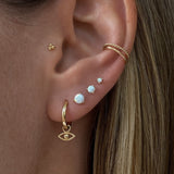 photo shows a close up of an ear with multiple earrings. wearing the golden Erin Huggie Hoop Earring in size Medium with the Jodi evil eye earring charm attached. the other earrings are 3 white opal studs in 3 different sizes, an ear cuff (Lani) and the Alma flat back piercing.