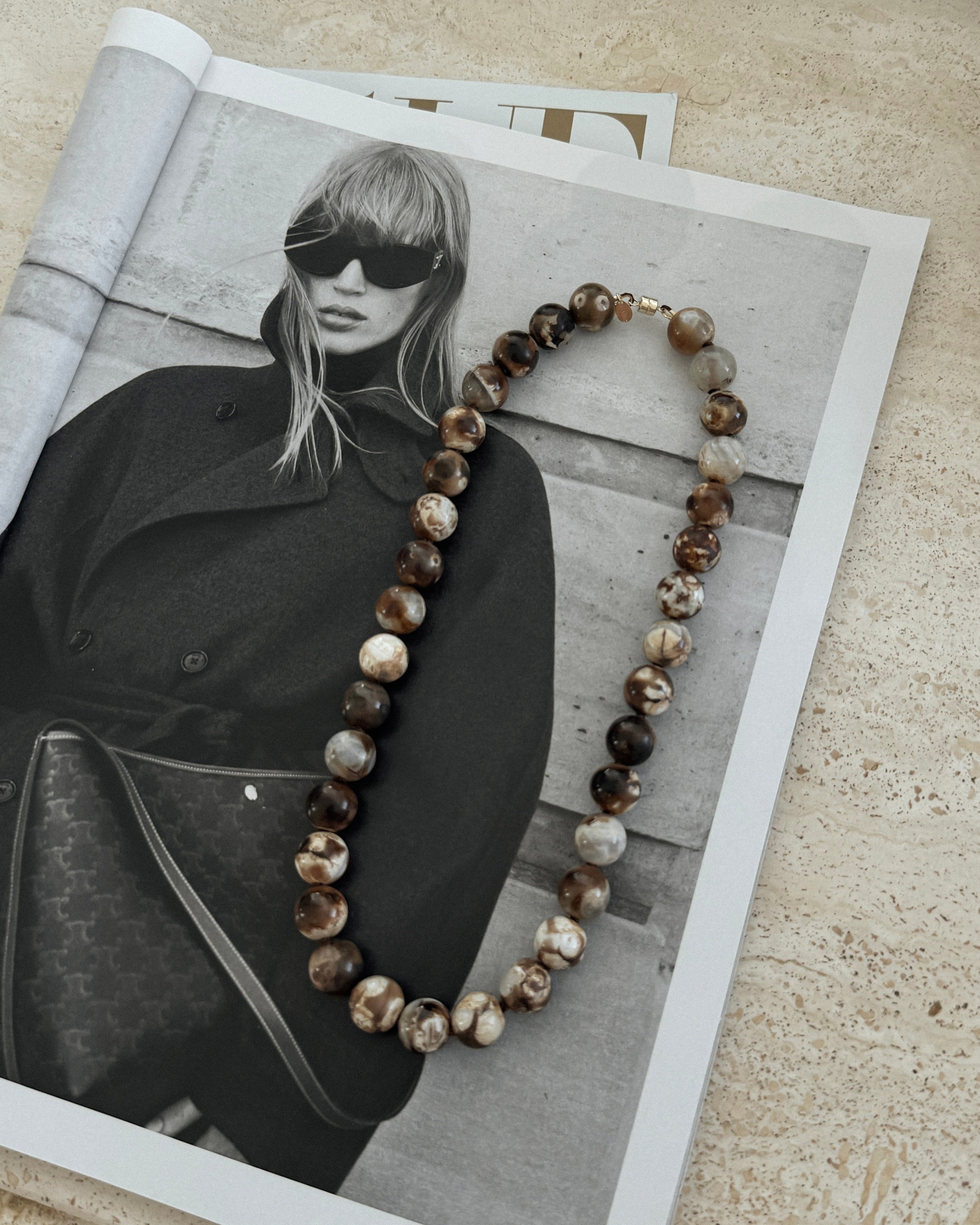 Brown beaded necklace on a magazine with a black and white photo of a woman wearing sunglasses and a coat.