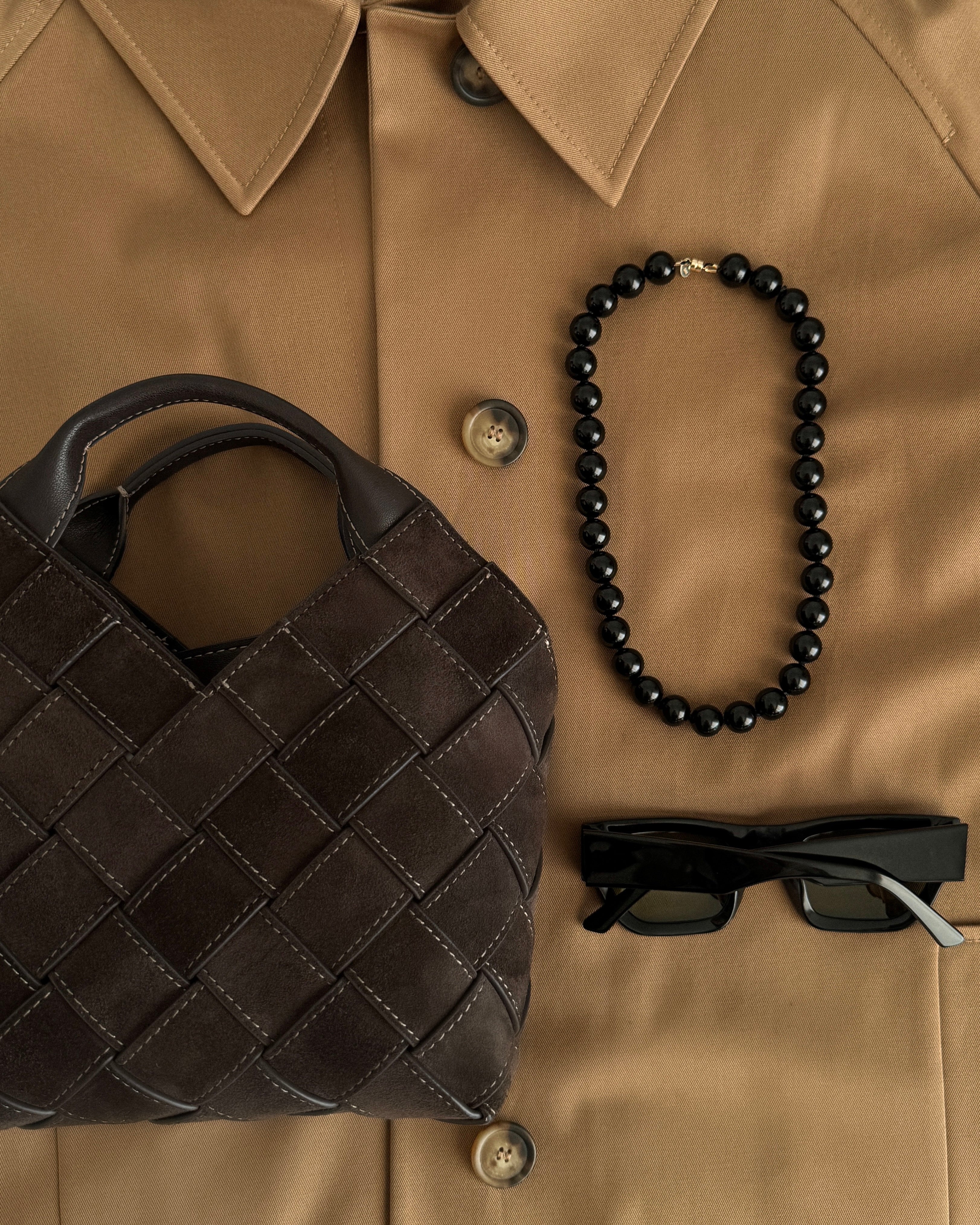 Brown quilted handbag, black beaded onyx necklace with magnetic clasp, and sunglasses on a beige coat.