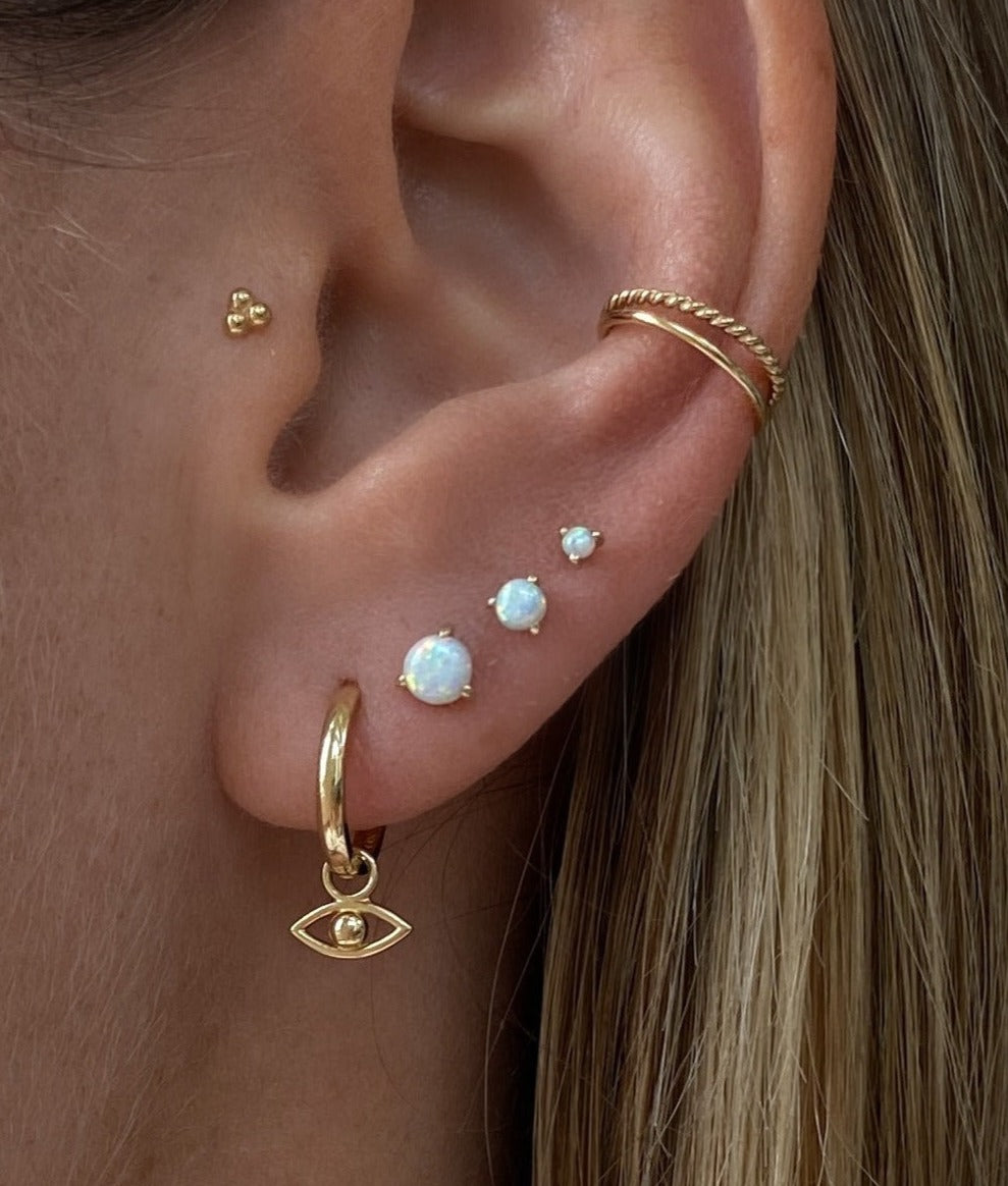 photo shows a close up of an ear with multiple earrings. wearing the golden Erin Huggie Hoop Earring in size Medium with the Jodi evil eye earring charm attached. the other earrings are 3 white opal studs in 3 different sizes, an ear cuff (Lani) and the Alma flat back piercing.